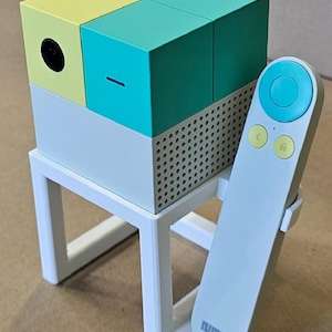 May include: A cube-shaped projector with yellow and teal panels, mounted on a white stand. A white remote control with teal accents rests beside it. The projector has a black lens and a series of small holes on the side.