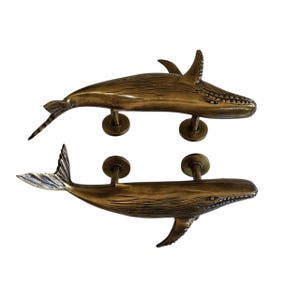 May include: Two bronze-colored whale-shaped door pulls. The whale pulls are detailed with fins, tails, and textured bodies. Each whale has two mounting posts. The door pulls are designed to be mounted horizontally.