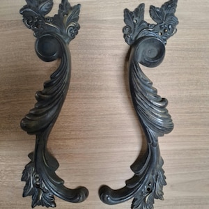 May include: Two ornate, dark bronze-colored door handles with a leaf and scroll design. The handles are curved with a detailed, textured surface. Small screws are visible below the handles.