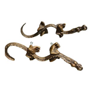 May include: Two bronze-colored door handles with an ornate design. The handles feature a curved shape with detailed, sculpted elements, including what appears to be a hand holding the handle and a decorative end.