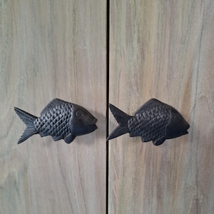 May include: Two dark gray, fish-shaped cabinet pulls mounted on a light-colored wooden cabinet door. The fish have detailed scales and fins, adding a decorative touch to the furniture. The cabinet doors have a light wood grain.