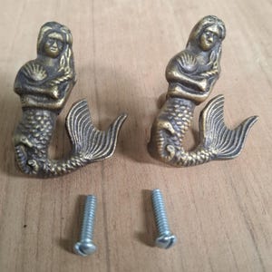 May include: Two gold-toned mermaid-shaped cabinet knobs with a detailed design. Each knob features a mermaid with a shell in her hand and a tail. The knobs come with two screws.
