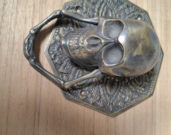 May include: A brass skull-shaped door knocker with a hexagonal backplate. The knocker features a detailed skull design and a ring-shaped handle. Includes four black screws.