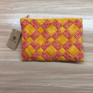 May include: A rectangular, yellow quilted pouch with a red and white diamond pattern. The pouch has a gold zipper and a small tag with the text "BATCH & STITCH". The pouch is lying on a light brown wooden surface.
