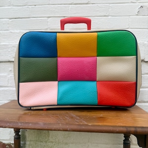 Puede incluir: Una maleta de patchwork multicolor con asa roja. La maleta rectangular presenta un borde color crema y un panel superior dividido en nueve cuadrados de varios colores, incluyendo azul, amarillo, verde, rosa y rojo. La maleta está sobre una mesa de madera.