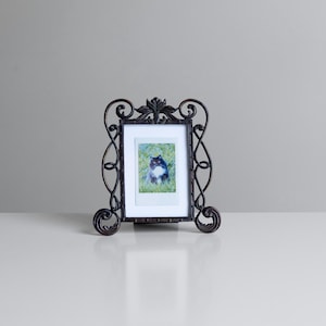 May include: A dark brown metal picture frame with an ornate, swirling scrollwork design and a central leaf motif at the top. The rectangular frame holds a small, vertical photograph of a black and white cat sitting in green grass.