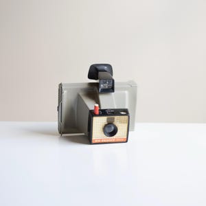 May include: A vintage Polaroid Big Swinger 3000 camera in a light gray color, with a black handle and flash. A smaller, black and gold camera is in front, with the text "BIG SWINGER 3000". The camera has a red button.