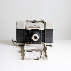 May include: A vintage gray and black camera with an open compartment revealing a paper insert. The camera has a large lens and a cord attached. The camera is on a white surface.