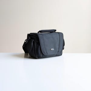 May include: A black camera bag with a structured rectangular shape, a top handle, and a front flap closure. The bag features a small Lowepro logo on the lower right corner of the front flap, side pockets, and durable fabric construction.