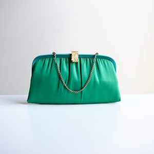 May include: Emerald green satin clutch with a gold-tone clasp and chain strap. The rectangular bag has a gathered design and a teal-colored frame. A classic evening bag.