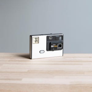 May include: A silver and black Kodak Disc 4000 camera. The camera has a rectangular shape with a flash on the left side and a lens on the right. The camera has a black section with the text "disc 4000".