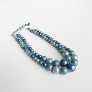 May include: A two-strand necklace featuring graduated blue beads in various shades. The necklace has a silver-tone clasp and is displayed on a white background. The beads range in size, creating a layered effect.