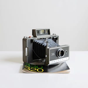 May include: A vintage Polaroid 330 Land Camera in gray and black, with a collapsible bellows and a prominent lens. The camera sits atop a stack of printed material with green and yellow text. The camera's design is classic and retro.