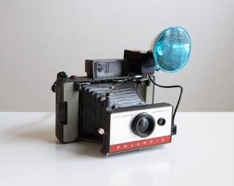 Puede incluir: Una cámara Polaroid Land Camera vintage, modelo Automatic 104, con cuerpo blanco y negro y un acento rojo. Cuenta con un flash azul y un fuelle negro. La cámara está sobre una superficie blanca.