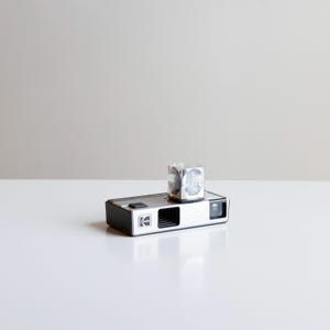 May include: A vintage Kodak camera with a silver and black body, featuring the Kodak logo. A silver cube-shaped flash attachment sits on top. The camera is on a white surface against a neutral background.