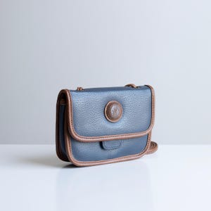 May include: A blue crossbody bag with brown trim and a circular emblem on the front. The bag has a textured appearance and a long strap. The bag is rectangular and has a flap closure.