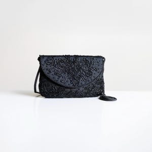 May include: Black beaded evening bag with a flap closure and a long, braided strap. The bag features an ornate, floral-inspired beaded design on the front. A small, beaded tassel hangs from the bottom right corner. The bag is set against a white background.
