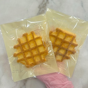 May include: Two individually packaged golden waffle cookies in clear plastic bags with yellow paper wrappers. The wrappers have a simple design with a small illustration of a cake and the text "not over 1 year".