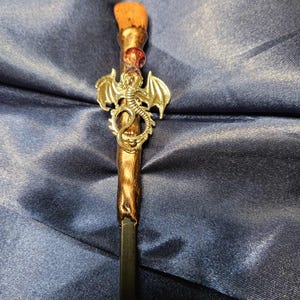 May include: A handcrafted wand with a silver dragon charm. The wand features a wooden handle with a brown and gold finish, a small red bead, and a light brown tip. The wand is a fantasy-themed item.