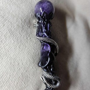 May include: A dark purple wand with a spherical amethyst crystal at the top. A silver-coloured snake wraps around the wand's body. The wand has a black base and intricate carved details.