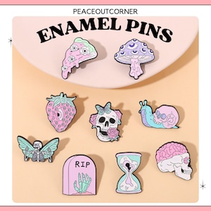 Könnte beinhalten: Set mit 10 Emaille-Pins mit verschiedenen Designs, darunter ein Pilz, eine Erdbeere, ein Schädel, eine Schnecke, ein Gehirn, eine Sanduhr, ein Pizzastück, ein Grabstein mit einer Hand und ein Skelett mit Flügeln. Die Pins sind alle in Pastellfarben gehalten und haben eine skurrile, gruselige Ästhetik.