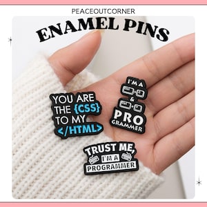Op de afbeelding: Vier emaille-pins met een coderingsmotief. De pins zijn zwart met witte tekst en blauwe accenten. De pins zeggen "You are the {CSS} to my </HTML>", "I'MA PRO GRAMMER", "Trust me, I'MA PROGRAMMER", en "Ctrl + C, Ctrl + V".