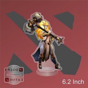 Puede incluir: Figura de acrílico de 15,7 cm de un personaje femenino del juego Dota 2, empuñando un gran martillo. La figura tiene pelo largo rubio y lleva una armadura en tonos blancos, plateados y dorados. El logo de Dota 2 es visible.