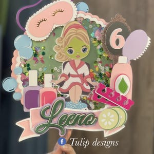 May include: A pink and green cake topper featuring a cartoon girl in a bathrobe and a face mask. The topper also includes the number 6, a nail file, nail polish bottles, and a cucumber. The text "Leena" and "Tulip designs" are also included.