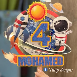 Puede incluir: Un adorno para pastel azul y naranja con un cohete, un astronauta, el sol, planetas y el número 4. El adorno también incluye el nombre "MOHAMED" y el texto "f Tulip designs".