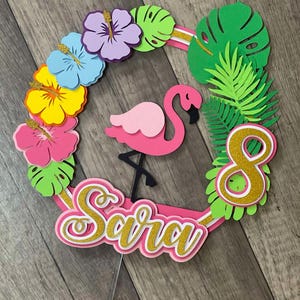 Op de afbeelding: Een roze flamingo taarttopper met de naam "Sara" in goudglitter schrift. Het ontwerp bevat een krans van kleurrijke papieren bloemen en groene bladeren, met het nummer "8" in goudglitter. De topper staat op een doorzichtige plastic stok.
