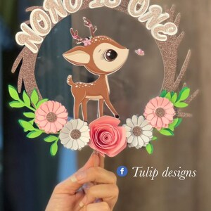 May include: A cake topper featuring a brown deer with pink flowers and greenery. The topper is decorated with glitter and the text "Nono is one".
