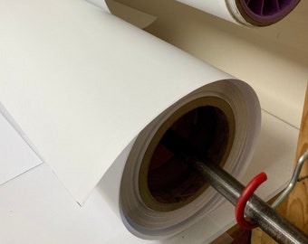 May include: Two rolls of white paper on a wooden surface. The rolls are on metal rods with a purple and black core on the top roll and a brown core on the bottom roll.