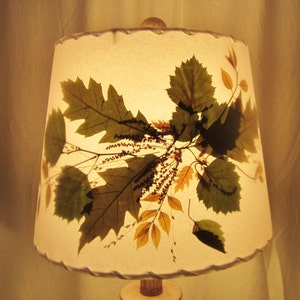 May include: A white lampshade with a pressed leaf design. The leaves are in shades of green, brown, and yellow. The lampshade has a scalloped edge.