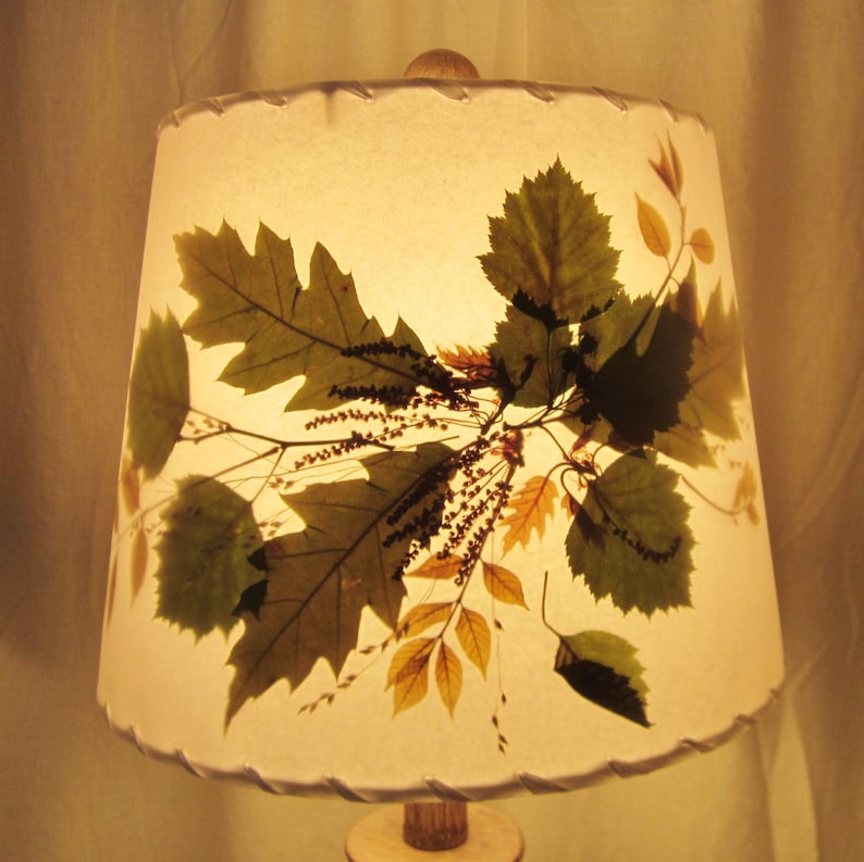 May include: A white lampshade with a pressed leaf design. The leaves are in shades of green, brown, and yellow. The lampshade has a scalloped edge.