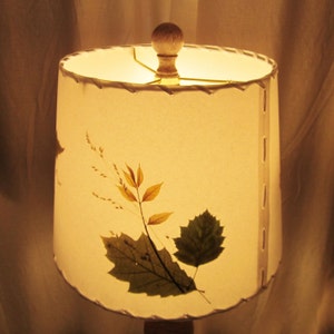 May include: A white lampshade with a pressed leaf design. The leaves are in shades of brown and green. The lampshade has a white fabric top and a white cord.