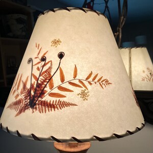 May include: A conical lampshade made of off-white, textured paper featuring pressed, dried fern fronds and small white flowers in shades of rust and brown. The top and bottom edges are finished with a dark brown, whip-stitched lacing detail.