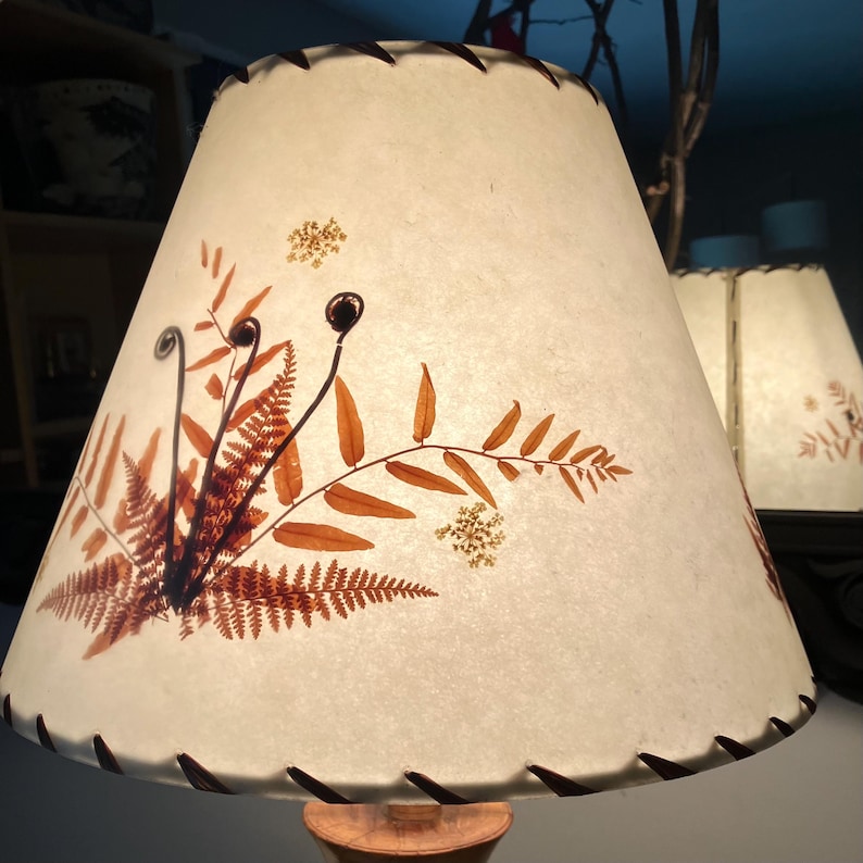 May include: A conical lampshade made of off-white, textured paper featuring pressed, dried fern fronds and small white flowers in shades of rust and brown. The top and bottom edges are finished with a dark brown, whip-stitched lacing detail.
