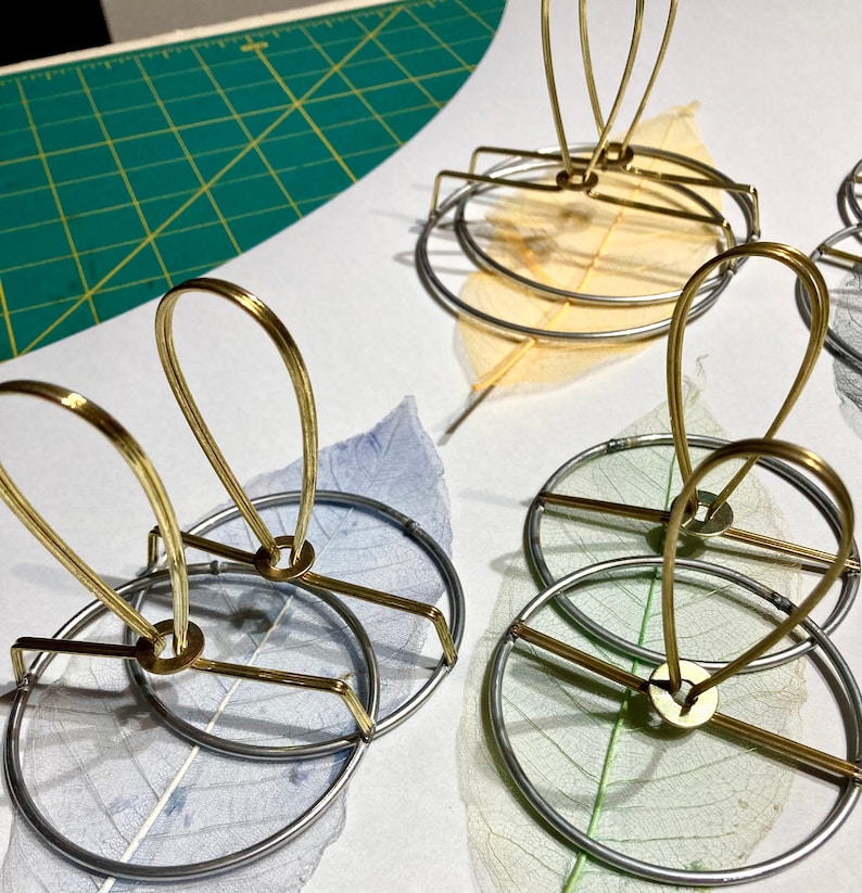 Puede incluir: Tres soportes de exhibici&oacute;n de metal dorado con delicadas hojas secas en tonos azules, verdes y amarillos. Los soportes presentan una base circular y una parte superior abierta curva.