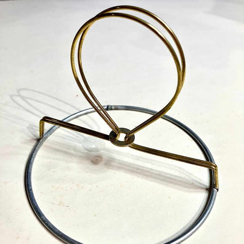 May include: A gold metal lampshade holder with a circular base and a ring at the top. The ring is double-layered and has a small metal piece in the center.