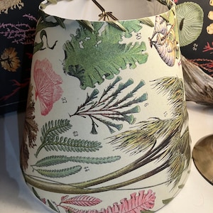 Könnte beinhalten: Ein weißer Lampenschirm mit einem botanischen Druck verschiedener Algen und Pflanzen. Der Druck ist in Grün-, Rosa- und Brauntönen gehalten. Der Lampenschirm hat einen braunen Besatz am oberen Rand.