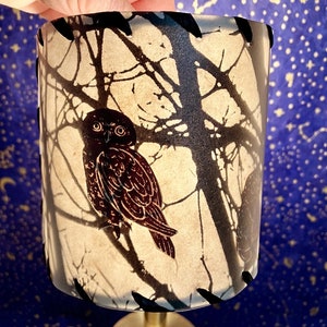 May include: A cream-colored lampshade with a black owl and tree branch design. The lampshade has a black stitched trim around the top and bottom.