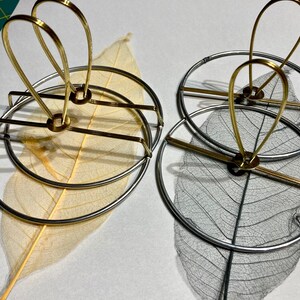 Puede incluir: Dos marcos circulares de metal con un delicado dise&ntilde;o de hoja en su interior. El marco de la izquierda es dorado y el marco de la derecha es plateado.