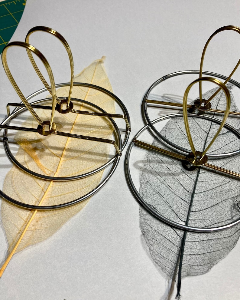 Puede incluir: Dos marcos circulares de metal con un delicado dise&ntilde;o de hoja en su interior. El marco de la izquierda es dorado y el marco de la derecha es plateado.
