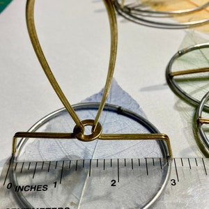 Puede incluir: Un soporte de metal dorado con una base redonda y un bucle en la parte superior. El soporte tiene un di&aacute;metro de 7,6 cm y est&aacute; sobre una regla.