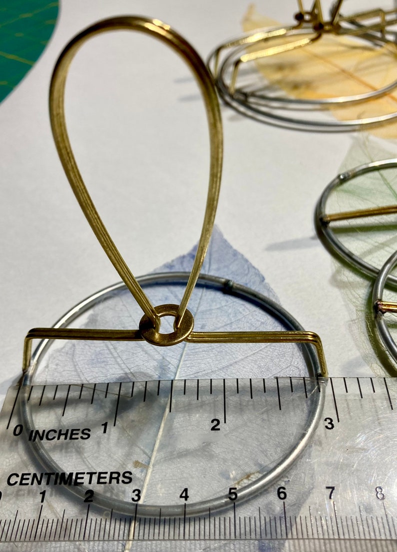Puede incluir: Un soporte de metal dorado con una base redonda y un bucle en la parte superior. El soporte tiene un di&aacute;metro de 7,6 cm y est&aacute; sobre una regla.
