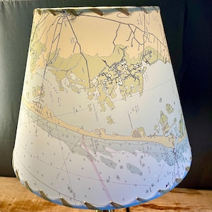 May include: A lampshade featuring a nautical chart design in shades of beige, green, and blue. The lampshade has a tapered shape and is trimmed with a decorative edge. The chart includes place names and depth markings.