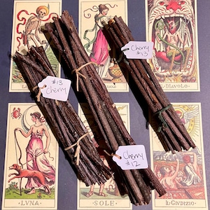 May include: Three bundles of brown sticks tied with twine, labelled "Cherry #13", "Cherry #12", and "Cherry #13". The bundles are arranged on a surface with colourful playing cards featuring images of skeletons, angels, and devils.