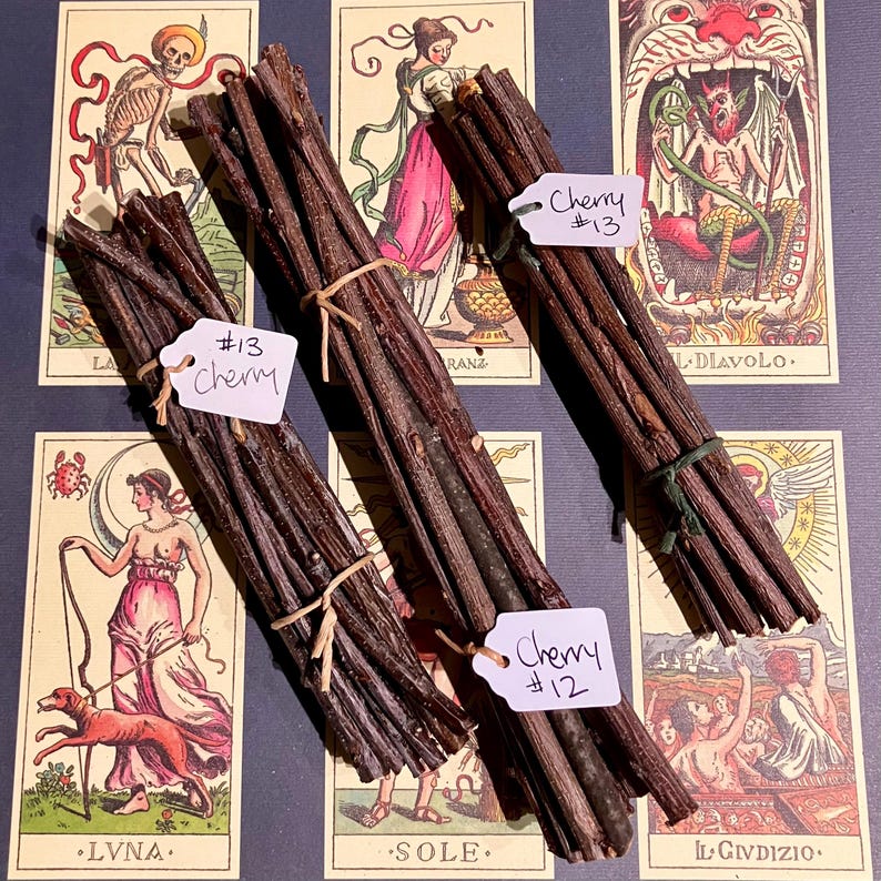 May include: Three bundles of brown sticks tied with twine, labelled "Cherry #13", "Cherry #12", and "Cherry #13". The bundles are arranged on a surface with colourful playing cards featuring images of skeletons, angels, and devils.
