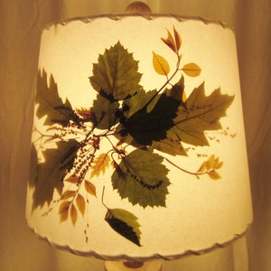 May include: A white lampshade with pressed leaves in shades of green, brown, and yellow. The leaves are arranged in a random pattern and create a natural, organic look.