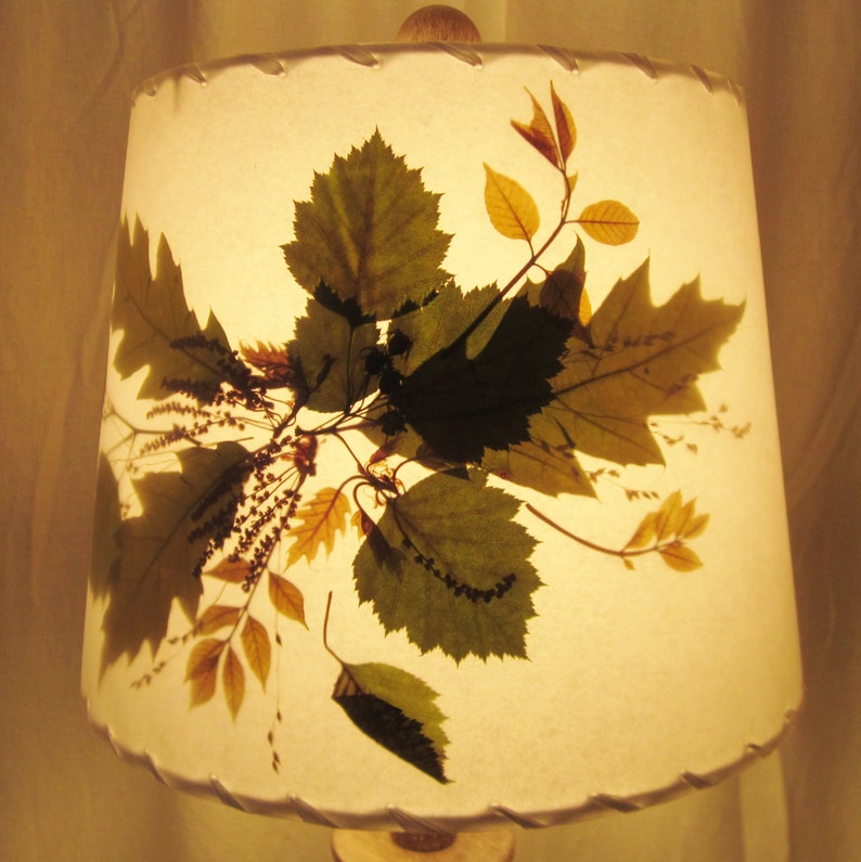 May include: A white lampshade with pressed leaves in shades of green, brown, and yellow. The leaves are arranged in a random pattern and create a natural, organic look.